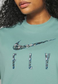 Valkoisen teepaidan, jossa on pyöreä kaula-aukko, vaalean teal-väri ja etupuolella on kuvioitu Nike-logon swoosh sekä sana "FLY" samantyyppisessä kuvioinnissa.