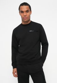 Felpa nera con scollo rotondo, realizzata in tessuto morbido. Presenta un sottile logo nero sul lato sinistro del petto. Fit rilassato con maniche lunghe.