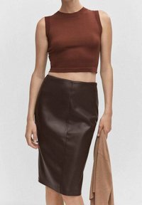 Un top court côtelé marron associé à une jupe crayon en cuir marron foncé, avec une texture lisse et une silhouette ajustée. Tenant un vêtement beige.