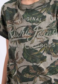 T-shirt à motif camouflage avec des feuilles vertes et des motifs floraux sur un fond gris. Il a un col rond et des manches courtes.