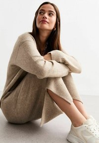 Beige gebreide jurk met ribbelaccenten en zijsplitten. Gecombineerd met lichte sneakers op een gladde grijze ondergrond.