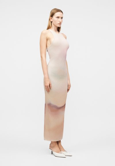 Γυναίκα ντυμένη με ένα αμάνικο, εφαρμοστό, μακρύ φόρεμα μπεζ χρώματος με απαλές μοβ αποχρώσεις, φορώντας λευκά μυτερά kitten heels, όρθια πλάγια.