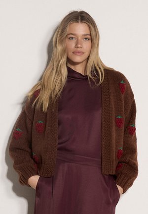 Frau mit langen blonden Haaren, die eine braune gestrickte Strickjacke mit roten Beeren über einem kastanienbraunen Kleid trägt, mit den Händen in den Taschen.