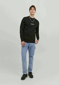 Young man standing wearing a black sweatshirt with white text, light blue jeans, black sneakers, and a silver chain necklace against a plain background.