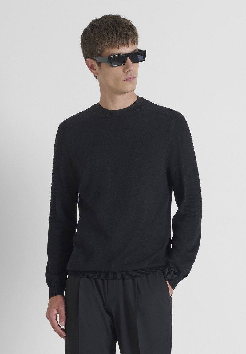 Maglione nero a girocollo con dettagli a coste sulle spalle, maniche lunghe e vestibilità rilassata, abbinato a pantaloni neri slim e occhiali da sole rettangolari.