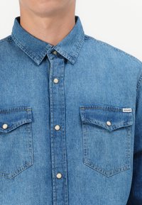 Camicia di denim azzurro chiaro con colletto classico, due tasche sul petto con chiusura a pressione e dettagli di cucitura discreti.