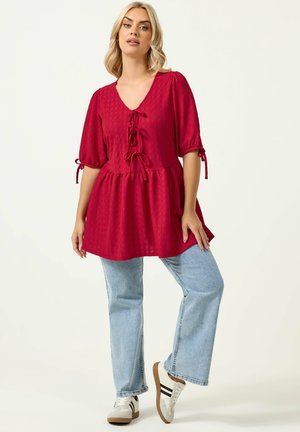 Lyshåret kvinde iført en rød tekstureret bluse med ballonærmer og bindebånd, lyseblå jeans og hvide sneakers med sorte striber.