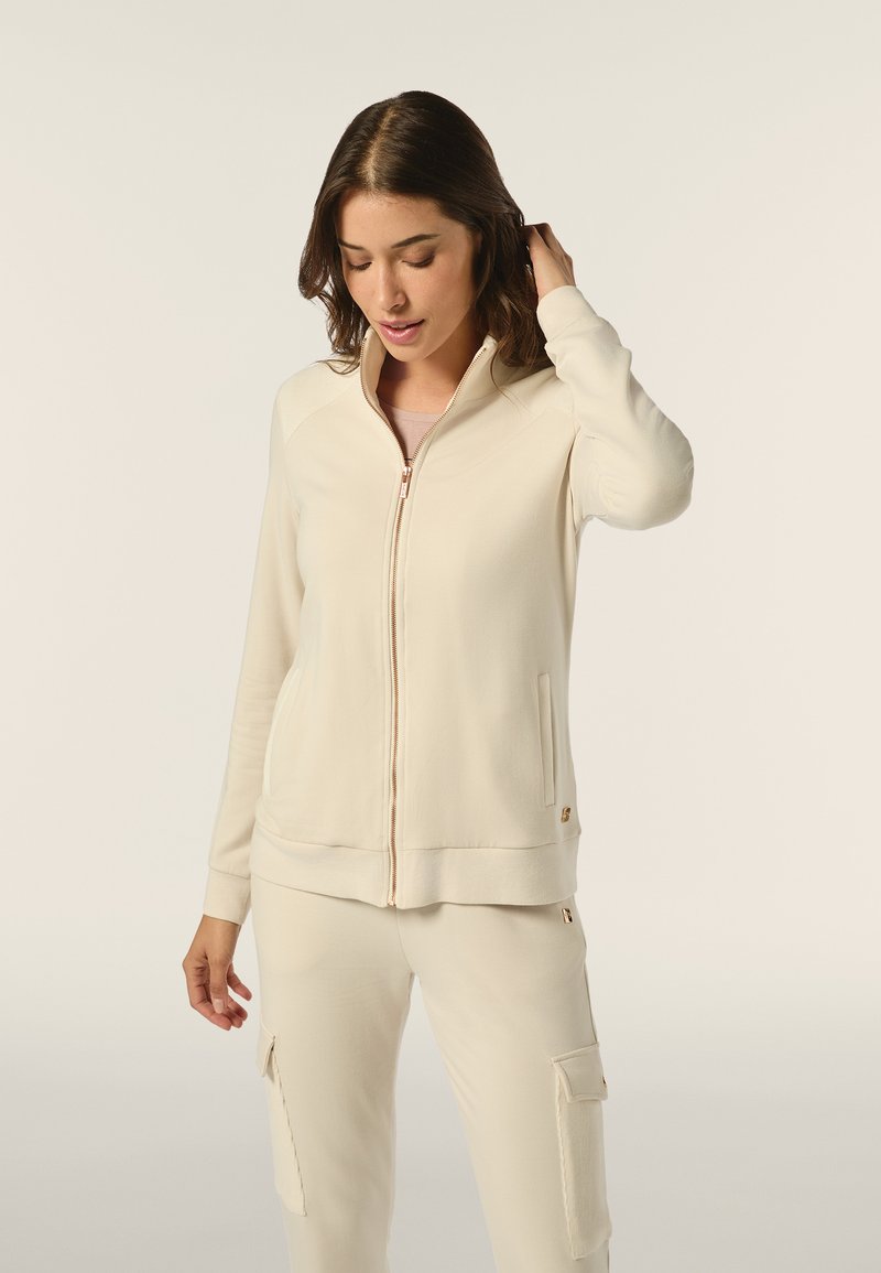 Veste zippée beige clair avec une texture lisse, des poignets côtelés et des poches latérales. Présente un col montant et des détails de fermeture éclair en ton doré.