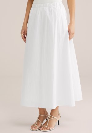 Donna che indossa una gonna bianca alla caviglia con vita elastica e sandali bianchi con tacco alto e cinturini, in piedi su uno sfondo neutro.