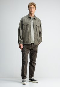 Jongeman in een olijfgroen jasje, lichtgrijze shirt, donkere cargobroek en zwarte sneakers, staande tegen een effen witte achtergrond.