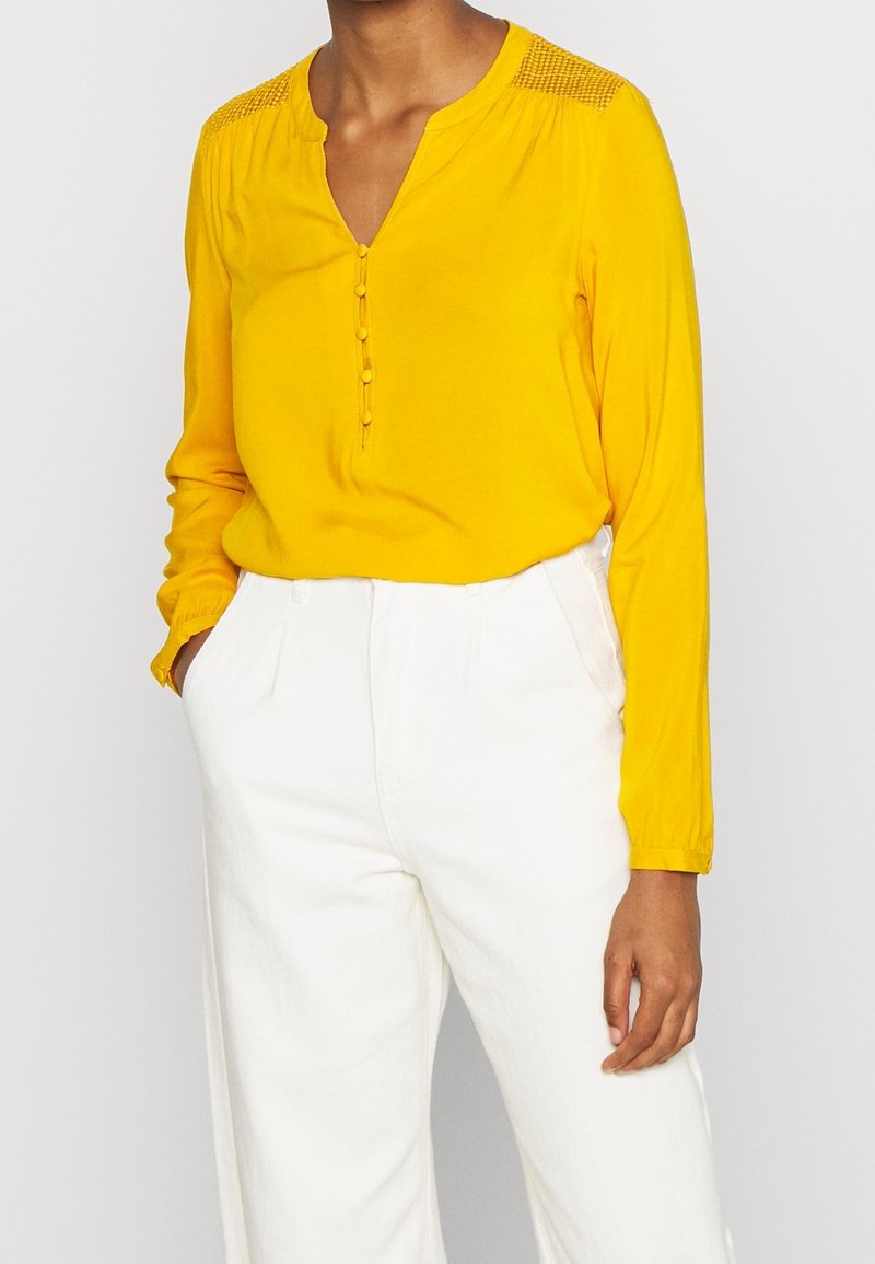 Personne portant un chemisier jaune à manches longues avec des boutons et un pantalon blanc taille haute, une main dans la poche, debout devant un fond uni.