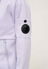 Veste violet clair avec fermeture éclair à l'avant, texture lisse et un accent noir rond sur la manche gauche avec un bouton de fermeture.