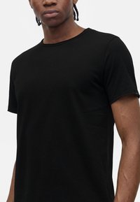 Jeune personne aux cheveux tressés portant un t-shirt noir uni à manches courtes sur un fond clair uni.