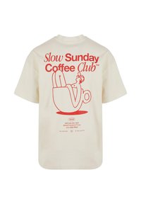 Bež majica z rdečim grafikonom, ki prikazuje osebo, ki počiva v skodelici kave, in besedilo "Slow Sunday Coffee Club" na hrbtu.