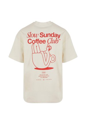 Bež majica s crvenom grafikom koja prikazuje osobu kako se opušta u šalici kave i tekstom "Slow Sunday Coffee Club" na leđima.