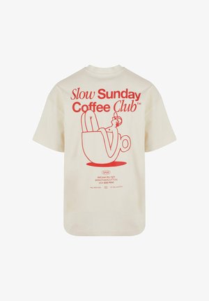 Smėlio spalvos marškinėliai su raudonu piešiniu, vaizduojančiu asmenį, atsipalaiduojantį kavos puodelyje, ir užrašas "Slow Sunday Coffee Club" nugarinėje dalyje.