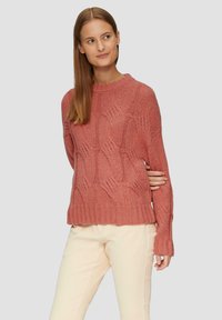 Sweter dzianinowy w miękkim różowym kolorze, z wzorem w liście, ribbowanym dołem i długimi rękawami. W zestawie z jasnobeżowymi spodniami.