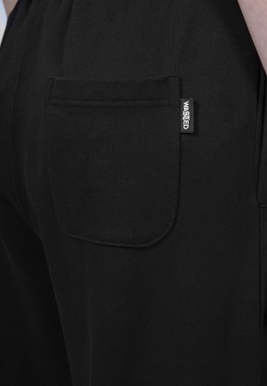 Gros plan sur la poche d'un pantalon noir avec une étiquette de marque "WASTED" sur la hanche d'une personne.