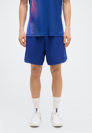 Muž v modrých športových šortkách a zodpovedajúcom športovom tričku, bielych ponožkách a bielych teniskách stojí na jednofarebnom pozadí.