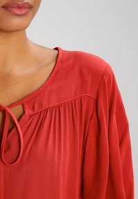 Blouse rouge avec un col rond et un détail de lien, en tissu texturé, et des plis doux à l'avant pour une dimension supplémentaire.