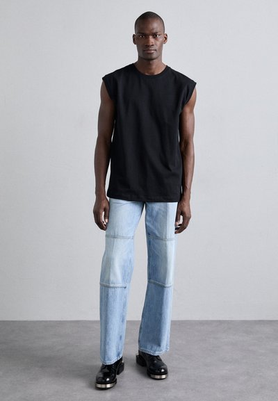 Tricou negru fără mâneci, blugi de culoare albastră deschisă cu panouri contrastante și pantofi platform negri, pe un fundal neutru.