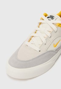 Nike SB tenisice imaju sivu i krem boju sa gornjim dijelom od velura i tekstila, bijelim vezicama i žutim akcentom loga sa strane.