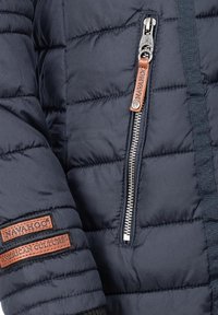Marinblå puffjacka med quiltat mönster, med en silverdragkedja och läderlogotyp på ärmen. Texturerad yta och slitstamt stoff.
