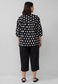 Schwarz-weißer Polka-Dot-Hemd mit Dreiviertelärmeln, rundem Ausschnitt, gerafftem Rücken, kombiniert mit knöchellangen schwarzen Hosen und schwarzen Sandalen.