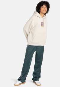 Crème-kleurige hoodie met een voorzak en abstracte grafiek; gecombineerd met donkergroene trainingsbroek met zijkanten en een relaxte pasvorm.