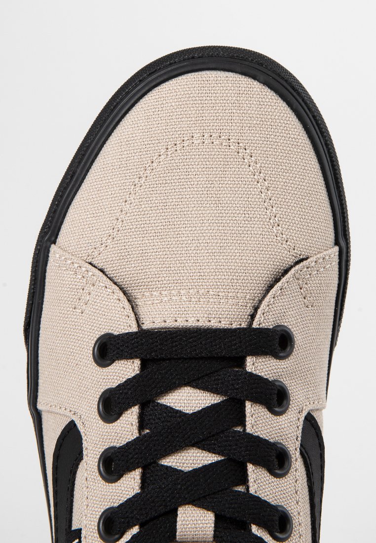 Baskets en toile beige avec des lacets noirs et un bord de semelle en caoutchouc noir, vues de dessus montrant les coutures détaillées et les œillets.