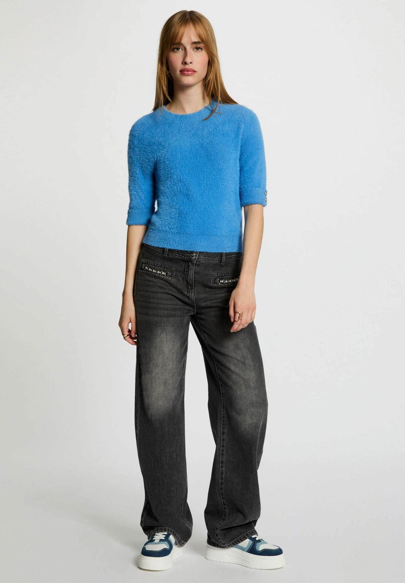 Maglione corto blu peloso con maniche corte, abbinato a jeans neri a gamba larga sbiaditi e accenti blu su sneakers bianche.