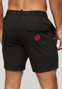 Zwarte zwemshorts met een elastische tailleband, achterzak en een rode logo-detail aan de rechterkant. Soepele stoftextuur.