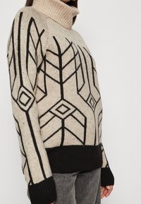 Beige stickad polotröja med svarta geometriska mönster, ribbad krage och mudd, med en avslappnad passform och texturerad tyg.