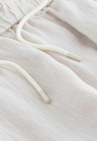 Gros plan sur un tissu beige clair avec des cordons blancs avec embouts en plastique aux extrémités.