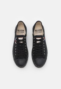 Baskets noires en toile avec embouts en caoutchouc, lacets ronds et accents jaunes. L'étiquette intérieure présente le nom de la marque et des détails sur son héritage.