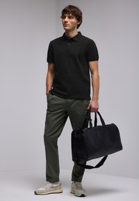 Svart pikétröja, gröna byxor och beige sneakers. Modellen håller en svart lädergympakväska med två handtag och en axelrem.