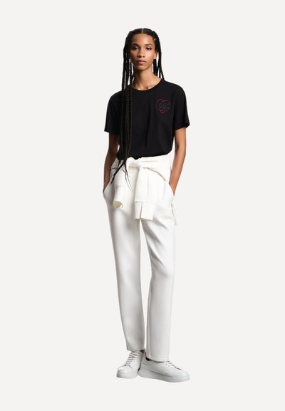 Giovane donna con trecce lunghe che indossa una maglietta nera con disegno a cuore, pantaloni bianchi, felpa bianca annodata in vita e sneakers bianche, in piedi.