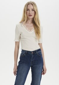 Kremasta rebrasta majica z V-izrezom in čipkastim robom, kombinirana z temno modrimi vračenimi jeans hlačami. Mehka tkanina z blagim raztezanjem.