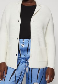 Mujer vestida con un cardigan blanco texturizado sobre una blusa negra y pantalones de satén azules con patrones abstractos en blanco y negro.
