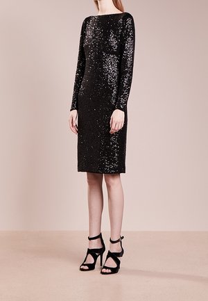 Femme debout portant une robe noire à sequins manches longues longueur genou et des sandales noires à talons hauts avec brides à la cheville.