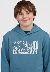 Smilende gutt med kort brunt hår, iført en blå O'Neill Santa Cruz hettegenser, mot en enkel hvit bakgrunn.