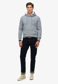Sudadera con capucha de color gris claro con un bolsillo frontal, combinada con jeans oscuros y zapatillas beige; presenta un estampado de logo en el pecho.
