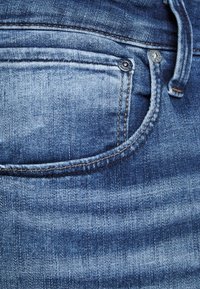 Blue denim jeans with a textured surface, featuring a side pocket, contrasting brown stitching, and a metal button on the waistband.