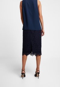 Top senza maniche blu navy abbinato a una gonna a matita in pizzo con orlo scallopato. La gonna presenta intricati disegni floreali e uno spacco posteriore. Tacchi alti neri.