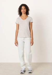 Mladá žena s kučeravými vlasmi stojí čelom, oblečená v svetlosivej tričku s výstrihom do V, bielych džínsoch a sivých teniskách.