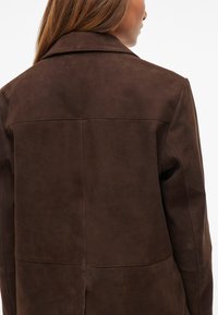 Casaco de camurça castanho com colarinho clássico, costuras visíveis e uma textura suave e matte. O design é ajustado com um corte estruturado.