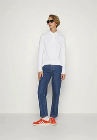 Jovem com óculos de sol pretos, polo de mangas compridas branco, jeans azuis, meias brancas e ténis Adidas laranja brilhante, de pé contra uma parede lisa.
