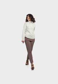 Hellgraue Zip-Jacke mit hohem Kragen, kombiniert mit braunen schmal geschnittenen Hosen und beigen Turnschuhen. Der Stoff wirkt glatt und flexibel.