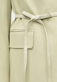 Gros plan sur une ceinture en tissu beige nouée en noeud papillon autour de la taille d'un manteau avec une poche à rabat sur le côté.