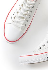 Zapatillas de lona blancas con acentos rojos, punteras de goma y cordones redondos. Suela texturizada con detalles de patrón antideslizante.